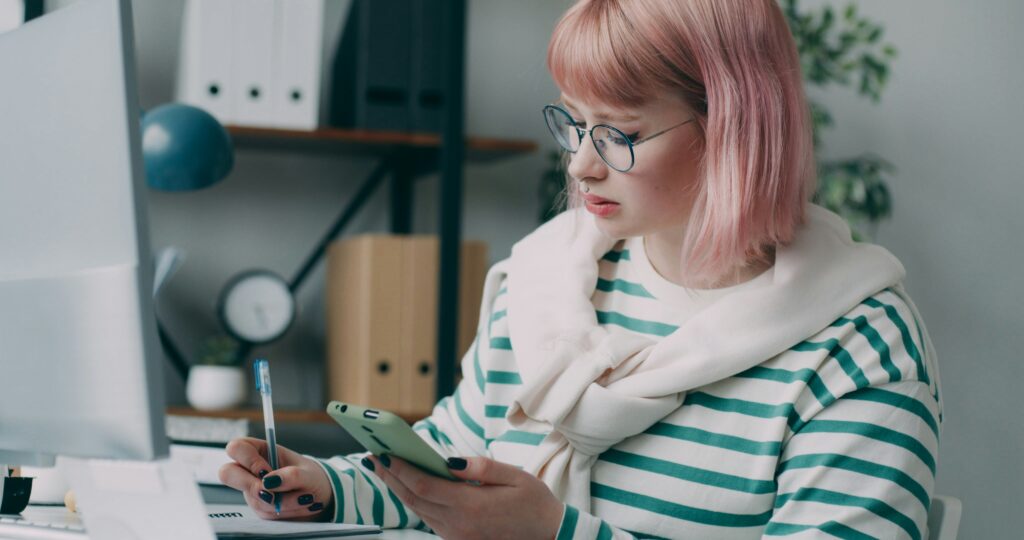 Uma mulher jovem, branca, e cabelo rosado, escreve em um caderno sobre mesa enquanto verificar a tela do celular com a outra mão. 