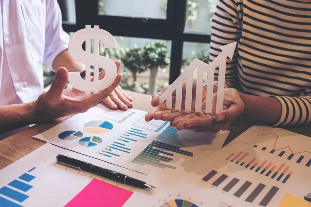 Duas pessoas analisam relatórios financeiros sobre uma mesa, segurando símbolos de cifrão e de crescimento, em referência a planejamento tributário e aumento de resultados.