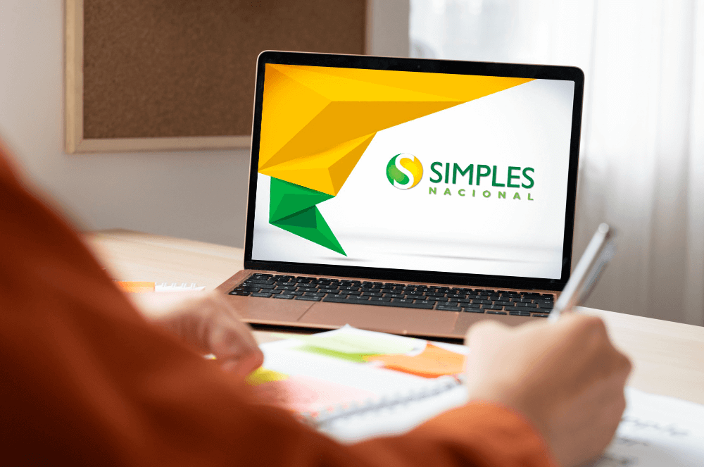 Pessoa anotando em um caderno diante de um notebook que exibe o logotipo do Simples Nacional.